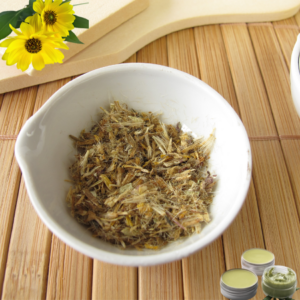 This image shows a bowl containing dried Arnica petals, set on a wooden surface. In the lower right corner, there are containers of green Arnica salve, indicating the use of the dried petals in making the salve. In the top left corner, there is an overlay of fresh Arnica flowers, visually connecting the dried petals and the salve to the live plant.