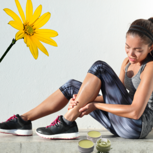 This image depicts a woman seated on the ground, holding her lower leg in apparent pain. She is dressed in athletic wear, including a grey tank top, grey and black gradient leggings, and black athletic shoes with pink laces. The expression on her face indicates discomfort. In the bottom corner, there are containers of a green salve, suggesting the use of a topical treatment. An Arnica flower is prominently displayed in the top left corner of the image, emphasizing its association with the salve for treating muscular injuries.