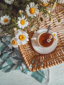 A cup of chamomile tea surrounded by fresh chamomile flowers on a wicker tray, with a soft teal cloth on a textured white surface.