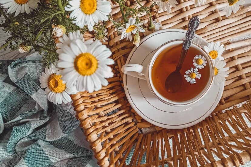 A cup of chamomile tea surrounded by fresh chamomile flowers on a wicker tray, with a soft teal cloth on a textured white surface.