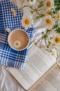 A cup of beverage with a chamomile flower on top, next to an open book and fresh chamomiles.