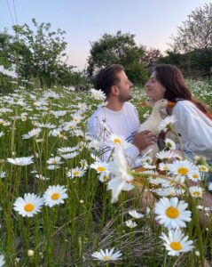 A couple sitting in a field full of blooming white pure camomile flowers, holding a duck, and gazing into each other's eyes.