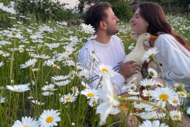 A couple sitting in a field full of blooming white pure camomile flowers, holding a duck, and gazing into each other's eyes.