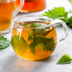 A clear glass cup filled with nettle tea surrounded by fresh nettle leaves on a light gray surface. Another glass of tea is partially visible in the background.