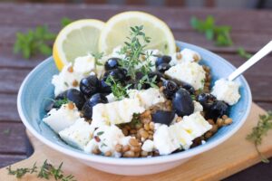 A bowl of lentil salad garnished with thyme, feta cheese, black olives, and lemon slices.