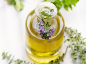 A clear glass bottle of golden thyme oil with a sprig of blooming thyme flowers inserted into its narrow neck, set against a white background with scattered thyme leaves and sprigs around it.