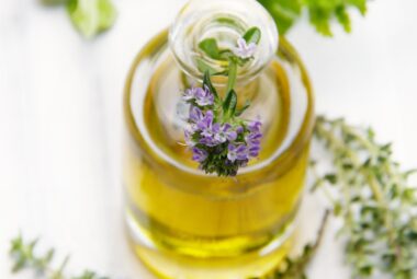 A clear glass bottle of golden thyme oil with a sprig of blooming thyme flowers inserted into its narrow neck, set against a white background with scattered thyme leaves and sprigs around it.