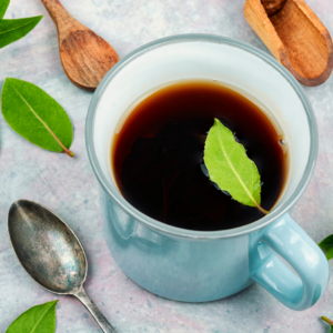 A blue cup filled with bay leaf tea, surrounded by fresh bay leaves, a wooden spoon, and a metal spoon on a light-colored surface.