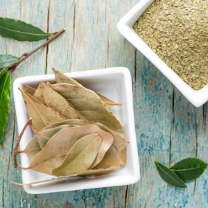  Dried bay leaves in a white dish, alongside ground bay leaf powder in another dish, with fresh bay leaves and a twig on a rustic wooden surface.