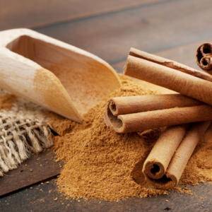 Article: How Can Cinnamon Control Blood Sugar Levels And Reduce Inflammation. A pile of ground cinnamon with a wooden scoop and several cinnamon sticks on a dark wooden surface.