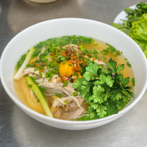 A bowl of chicken noodle soup garnished with fresh herbs, featuring a prominent coriander illustration on the side.