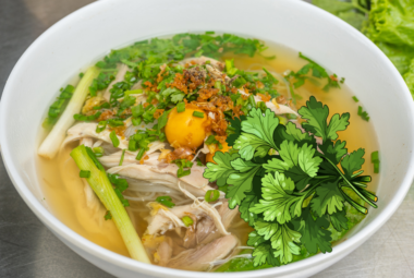 A bowl of chicken noodle soup garnished with fresh herbs, featuring a prominent coriander illustration on the side.