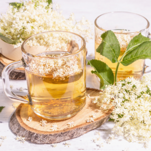 Article: Immune Boosting Elderberry Tea Benefits. Pic "Two glass mugs of elderflower tea with fresh elderflowers and green leaves on a white wooden surface."