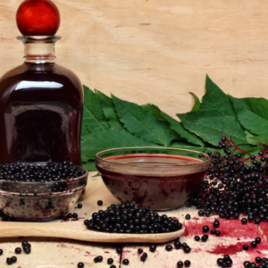 Article: Immune Boosting Elderberry Tea Benefits. Pic "Glass bottle and bowl of elderberry juice, fresh elderberries, and green leaves on a wooden surface."