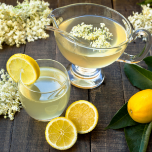 "Glass pitcher and glass of elderflower lemonade garnished with lemon slices and elderflowers, with lemons and leaves on a wooden table."