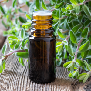 A small amber glass bottle with a white cap, placed on a wooden surface surrounded by fresh marjoram leaves.