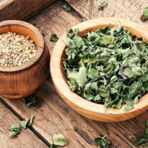  Two wooden bowls on a rustic wooden surface, one filled with dried marjoram leaves and the other with a blend of dried herbs.