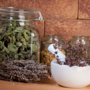 A collection of dried herbs in glass jars, with a mortar and pestle filled with dried marjoram flowers, set against a rustic brick background.