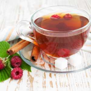 Article: Raspberry Leaf tea. Pic -A glass cup of raspberry leaf tea with cinnamon sticks, fresh raspberries, and sugar cubes on a wooden table.