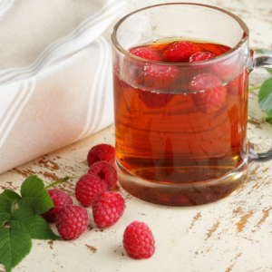  A glass mug filled with raspberry leaf tea, fresh raspberries floating on top, set on a rustic painted table with loose raspberries and leaves around.