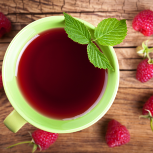 Overhead view of a cup of raspberry leaf tea in a green mug, garnished with fresh raspberry leaves, on a wooden table surrounded by raspberries.