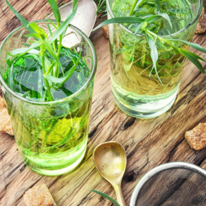  Two glasses of tarragon tea with fresh tarragon leaves, surrounded by a spoon, sugar cubes, and a tea strainer on a wooden surface.