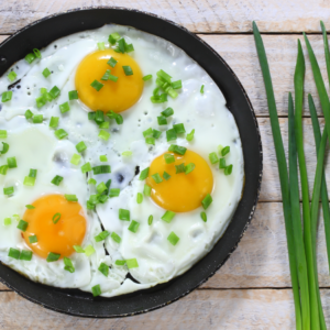  A top view of three sunny-side-up eggs in a frying pan, garnished with chopped chives, with fresh chive stalks beside the pan.