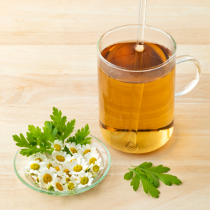  A glass cup of herbal tea with a strainer, accompanied by a small dish of feverfew flowers and leaves on a wooden surface.