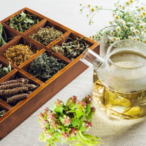 : A glass teapot with herbal tea next to a wooden box of dried herbs and a bunch of fresh herbs, with chamomile flowers in the background.