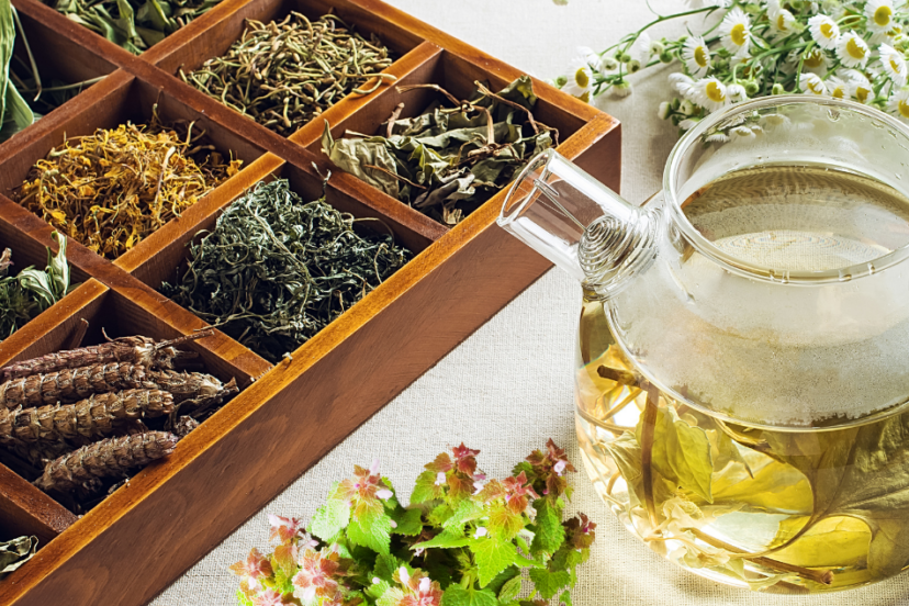 : A glass teapot with herbal tea next to a wooden box of dried herbs and a bunch of fresh herbs, with chamomile flowers in the background.