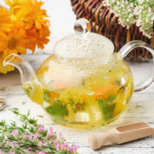 A clear glass teapot filled with freshly brewed herbal tea, surrounded by vibrant flowers and herbs on a rustic white table.