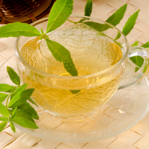 A clear glass cup filled with herbal tea, garnished with fresh lemon verbena leaves, placed on a matching glass saucer.