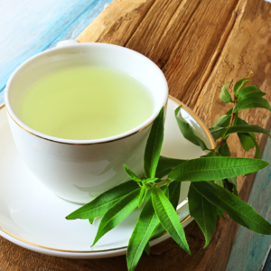 A white ceramic cup filled with light green herbal tea, garnished with fresh lemon verbena leaves, placed on a rustic wooden surface.