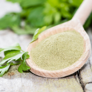 Close-up of a wooden spoon filled with ground lovage powder, with fresh green lovage leaves in the background.