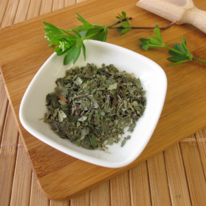 Dried woodruff leaves in a white bowl on a wooden board with fresh woodruff sprigs beside it.