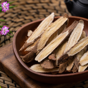 Article: Astragalus tea. Pic - A wooden bowl filled with dried astragalus root slices, placed on a woven mat surrounded by small pink astragalus flowers