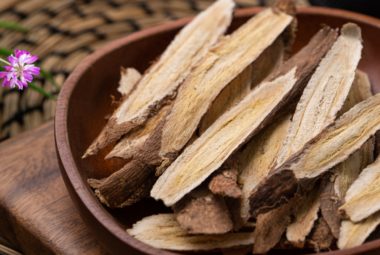 A wooden bowl filled with dried astragalus root slices, placed on a woven mat surrounded by small pink astragalus flowers