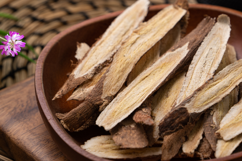 A wooden bowl filled with dried astragalus root slices, placed on a woven mat surrounded by small pink astragalus flowers