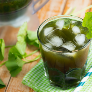  Glass of green gotu kola juice with ice cubes and fresh leaves on a wooden table.