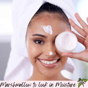  A smiling person with a towel on their head applies marshmallow root cream to their face while holding a jar.