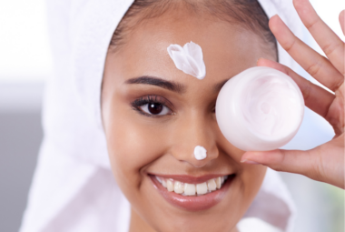 A smiling person with a towel on their head applies marshmallow root cream to their face while holding a jar.