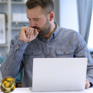 man sitting at a desk with a laptop, coughing into his hand, with a cup of coltsfoot tea nearby.