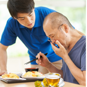 an older man with a meal, as the older man holds his hand to his mouth coughing, with a cup of herbal tea and coltsfoot flowers on the table.